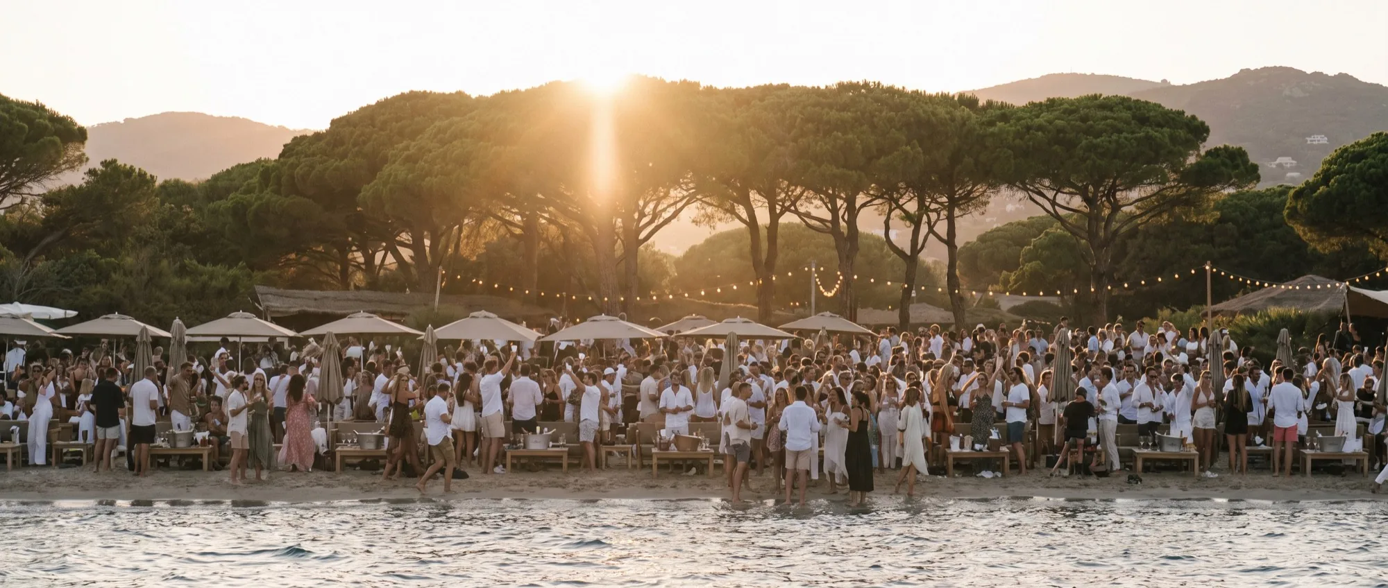 Un beach club de la Côte d’Azur en pleine fête à l’heure dorée, les invités dansant sur le sable, coupe à la main, sous les guirlandes.